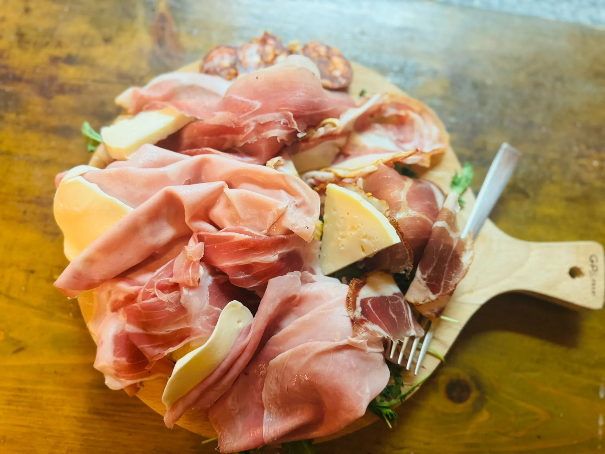 Tagliere di legno con prosciutto crudo, salame, spicchi di formaggio, rucola e forchetta: antipasto de Il Quartino