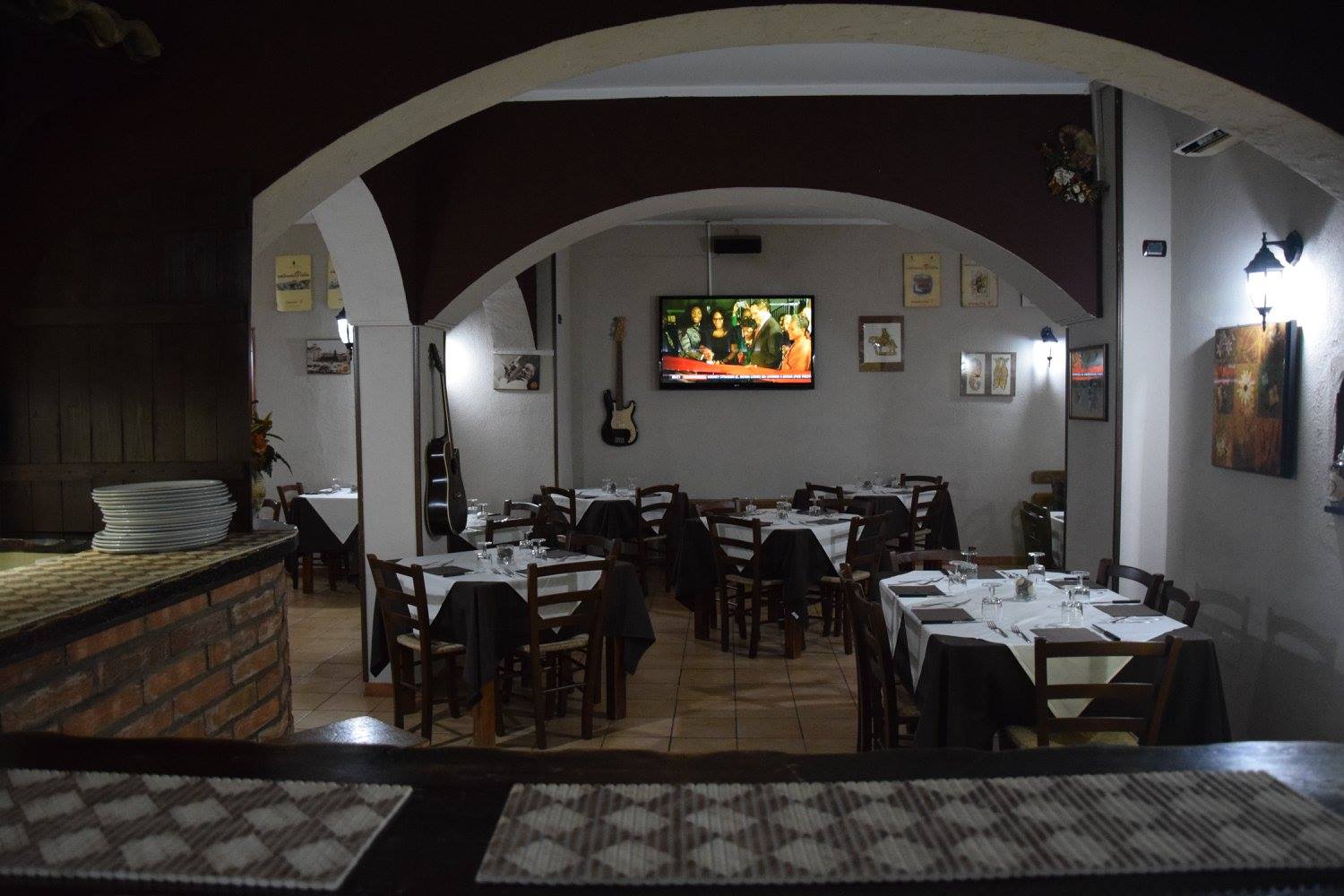 Sala del Quartino con archi bianchi, tavoli con tovaglie nere e bianche, sedie in legno, chitarre appese e TV a parete